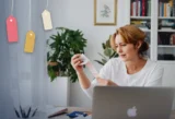 Small business owner reviewing receipts at her laptop with a calculator, with price tags hanging beside her